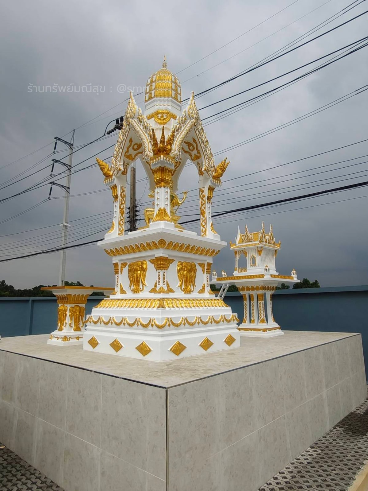 ศาลพระพรหมทรงไทย PST-0020 สีขาวตัดทอง มุมเฉียงบนแท่นยกสูง เห็นจั่วแหลม ใบระกา และลายช้างที่ฐาน