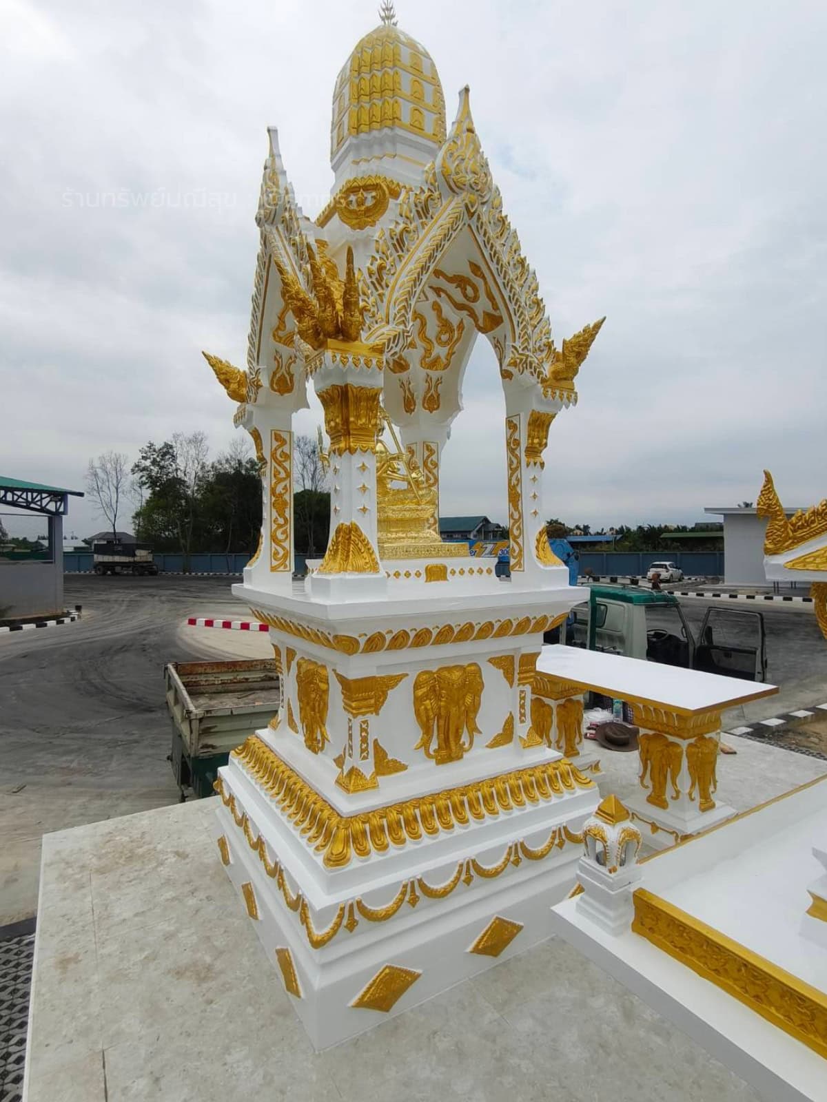 ศาลพระพรหมทรงไทย PST-0020 สีขาวตัดทอง มุมเฉียงระยะใกล้ เห็นซุ้มจั่วแหลม เสา ลายช้าง และโต๊ะด้านข้าง